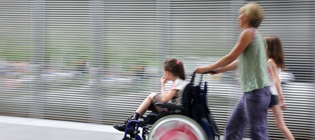Ein Mädchen in Begleitung ihrer Schwester wird von der Mutter im Rollstuhl geschoben.