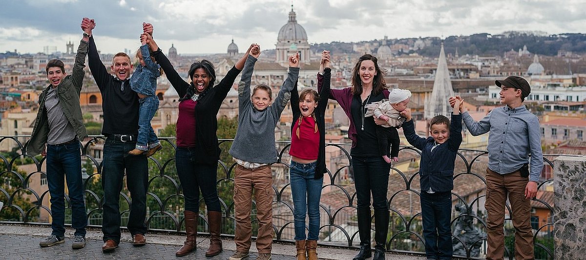 Die Mitglieder einer großen, bunten Familie halten sich in Rom bei den Händen und heben euphorisch die Arme.