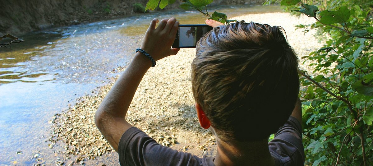 Ein Junge steht vor einem Fluss und fotografiert diesen mit dem Smartphone