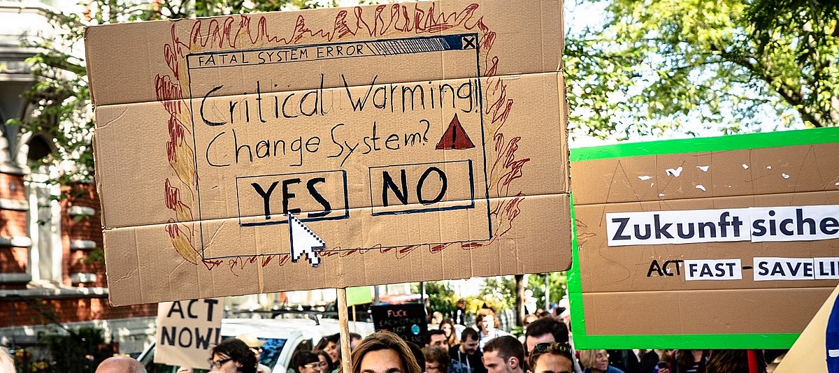 Ein Protestzug junger Menschen, im Zentrum ein Schild mit der Aufschrift Critical Warming - Change System? und einem Pfeil, der auf Yes deutet