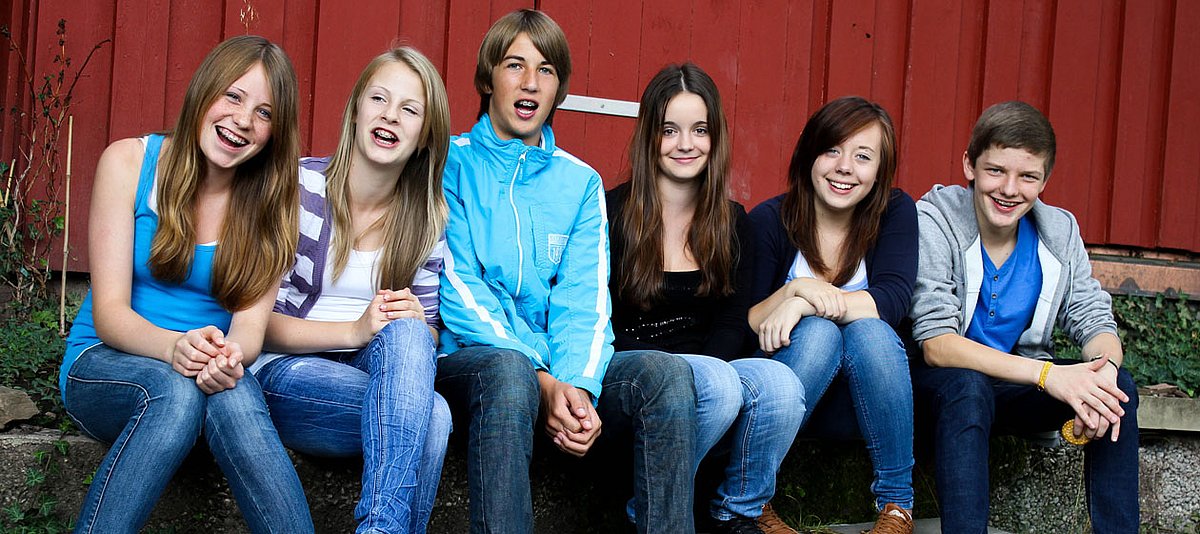 Eine Gruppe Jugendlicher sitzen vor einer roten Holzwand und lachen in die Kamera. 