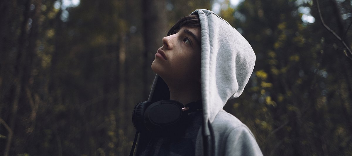 Ein Junge mit Kapuzenpulli und umgehängten Kopfhörern steht im Wald und schaut hoffnungsvoll nach oben