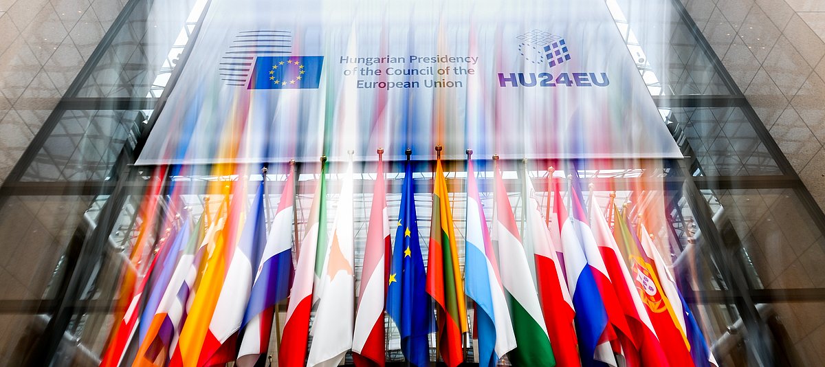 "Hungarian presidency of the Council of the European Union" steht als Text auf einem großen Banner. Davor hängen europäische Flaggen.