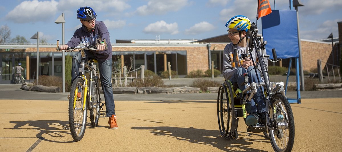 Jugendlicher mit Behinderung sitzt in seinem Fahrrad und schaut zu einem Jugendlichen ohne Behinderung, ebenfalls auf seinem Fahrrad