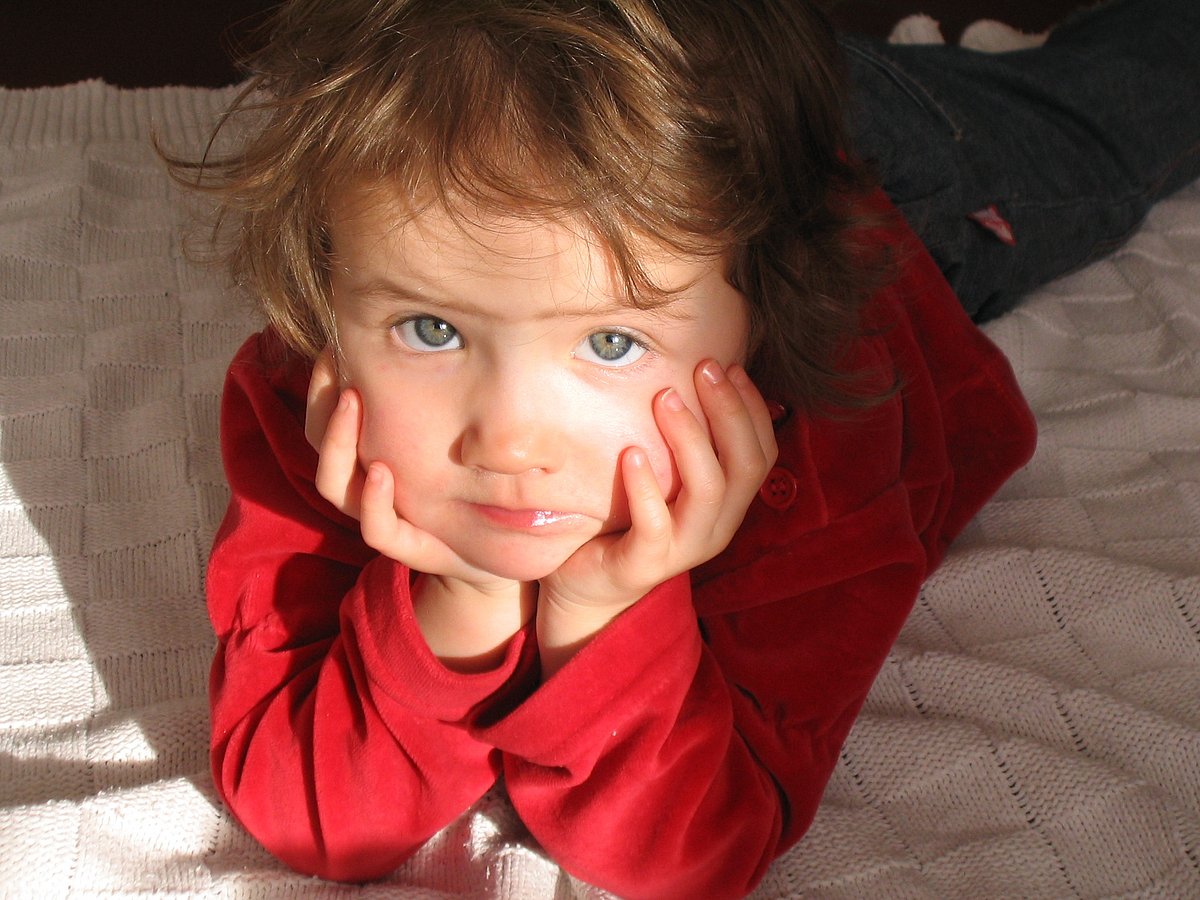 Ein blondes Mädchen liegt auf einer Decke und stützt den Kopf auf die Hände. Es trägt einen roten Pullover und schaut traurig in die Kamera. 