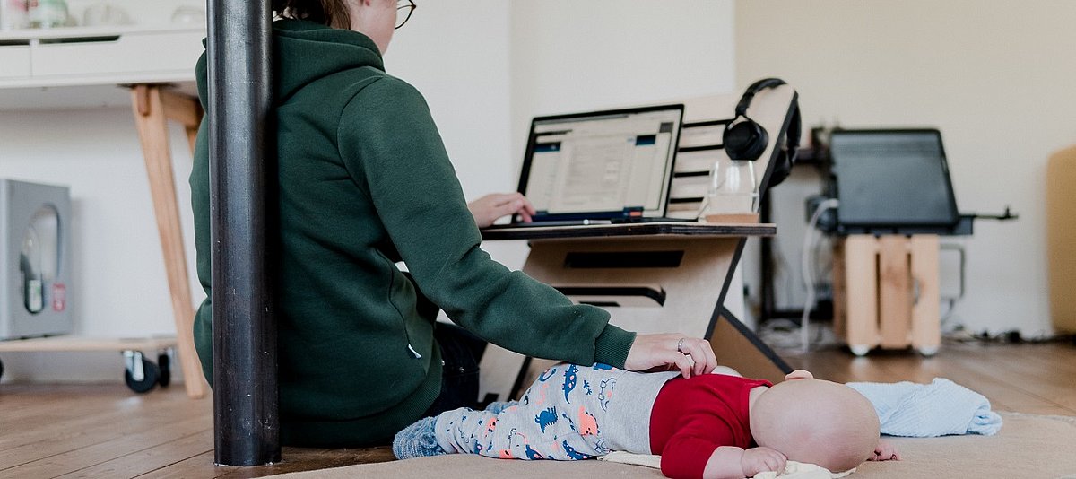 Eine Mutter, die das neben sich liegende Baby streichelt, arbeitet von zuhause an einem Laptop