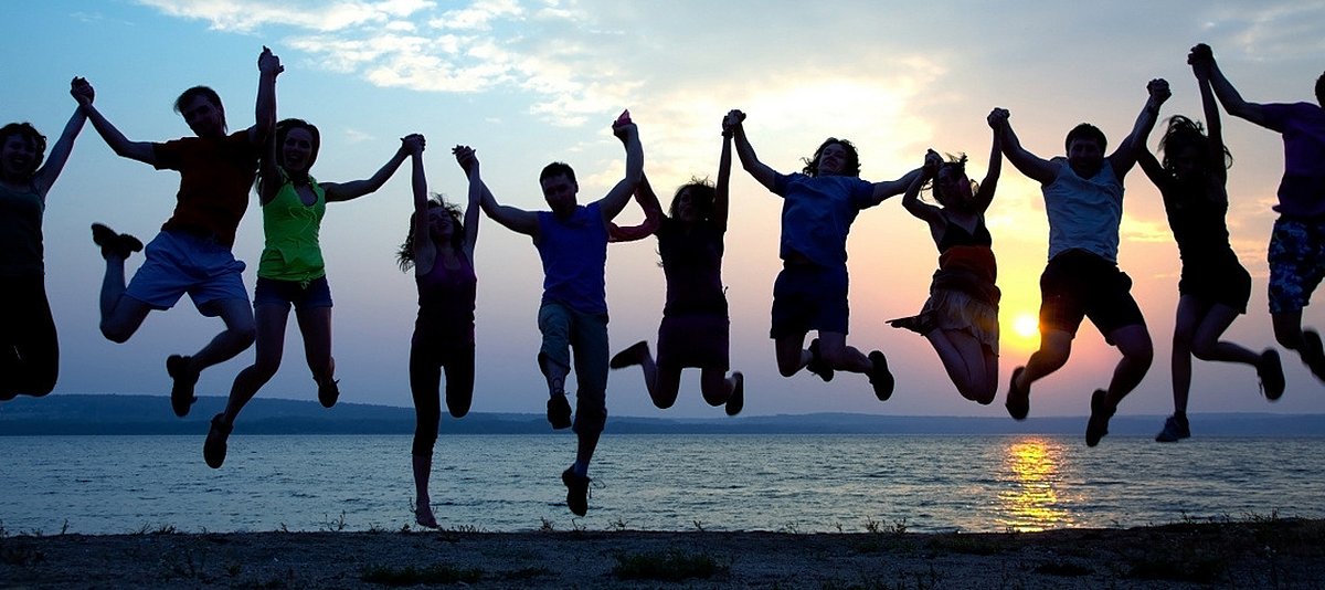 Junge Menschen halten sich an den Händen und springen in die Luft. Im Hintergrund sieht man die untergehende Sonne über einem See. 