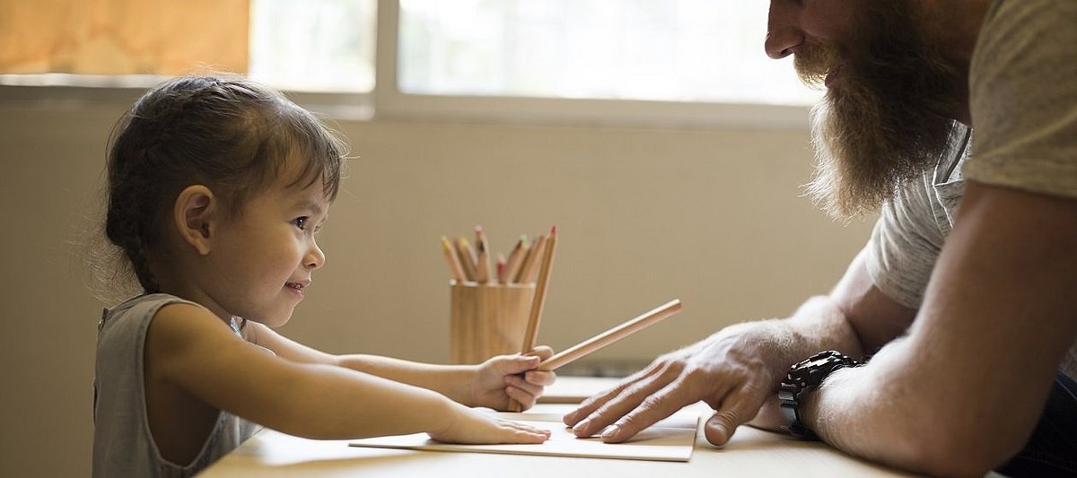 Ein Kind sitzt an einem Tisch und hat Stifte in der Hand, gegenüber sitzt ein Erwachsener
