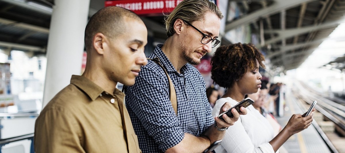Drei Menschen stehen nebeneinander an einem Bahngleis und schauen auf ihr Smartphone