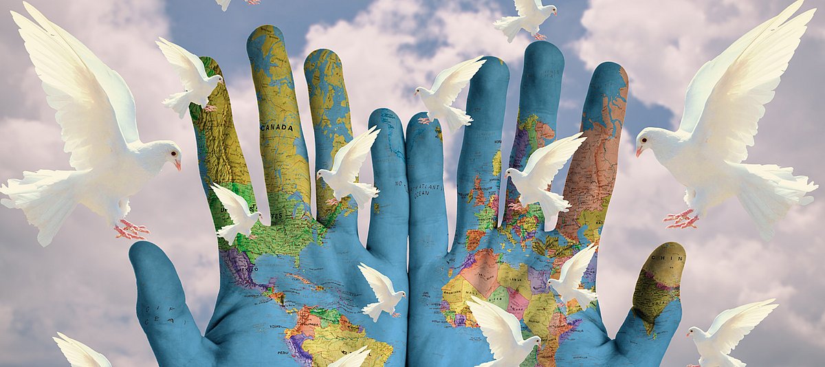 Eine Weltkarte in zwei Hände gezeichnet vor blau-weißem Himmel und weißen Friedenstauben. 
