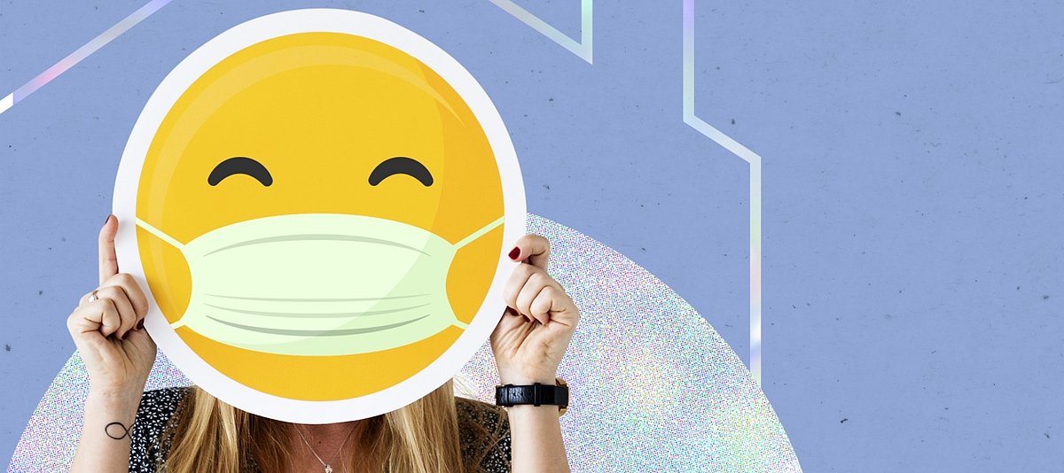 Person hält ein Schild eines Emoji mit Mundschutz vor ihren Kopf