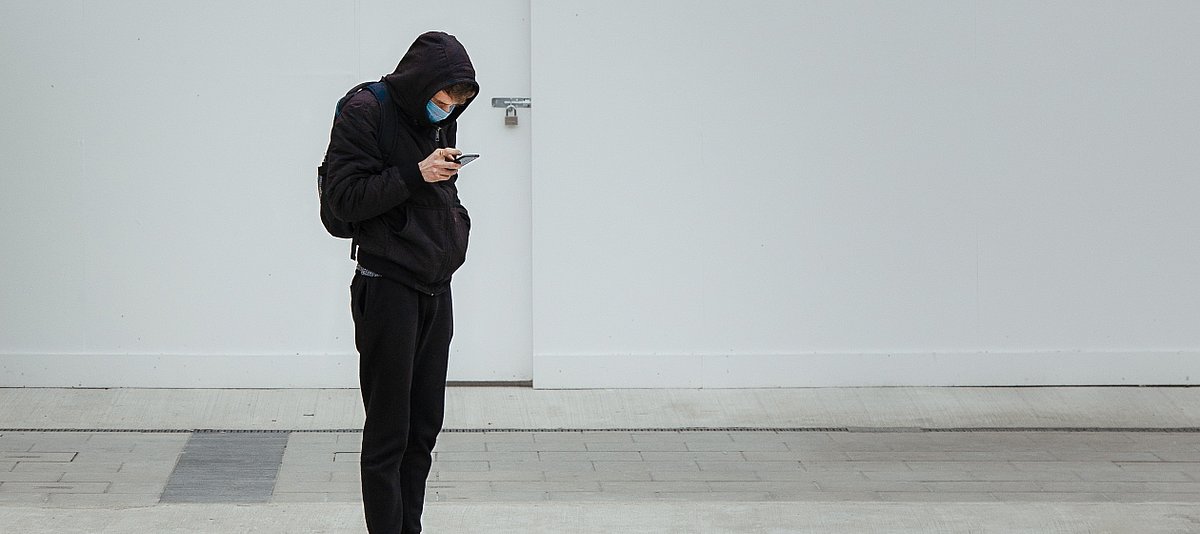 Ein Jugendlicher in schwarzer Kleidung trägt einen Mundschutz und schaut auf sein Smartphone