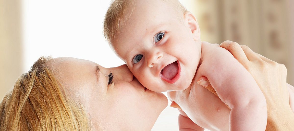 Eine Frau hält ihr Baby hoch und küsst es. Das Baby lacht dabei.