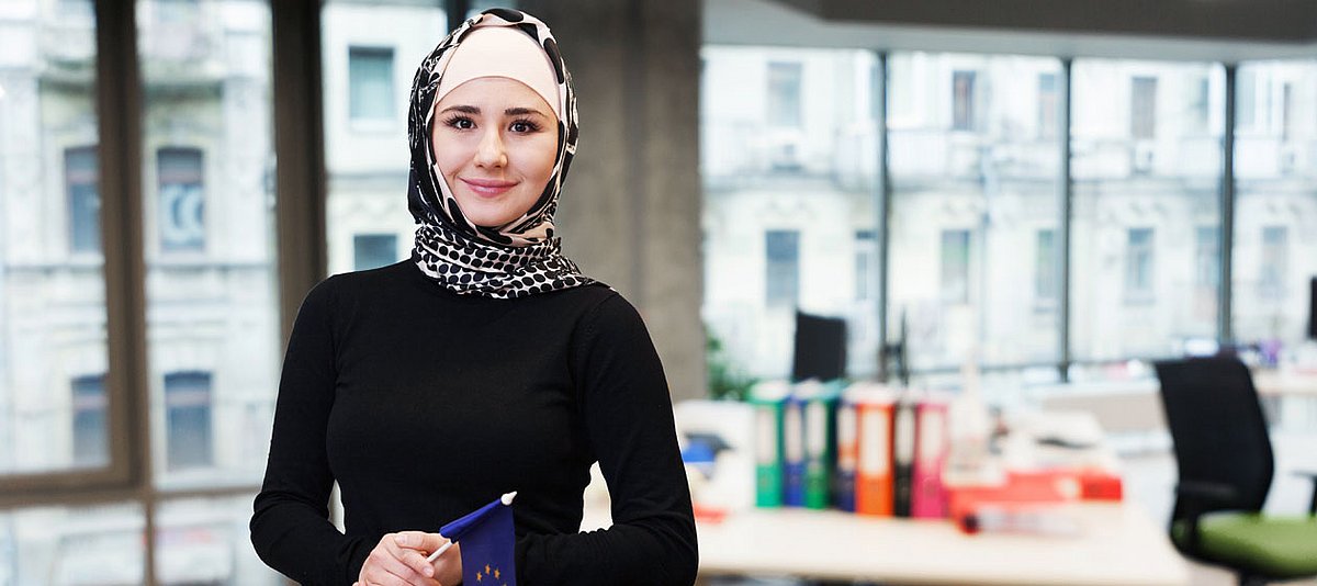 Junge Frau mit Kopftuch und einer EU-Flagge in der Hand steht in einem Büro und lächelt. 