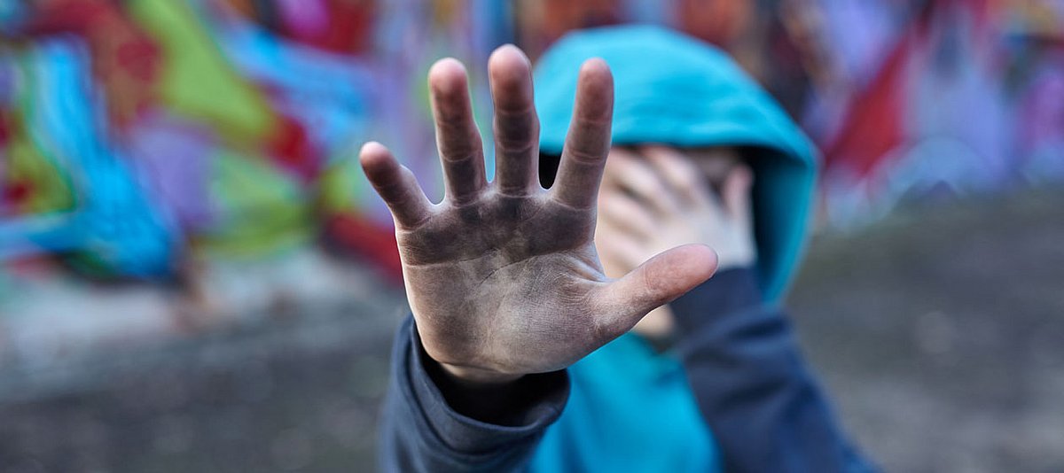 Ein Teenager steht vor einem Graffiti und hält seine schmutzige Hand abwehrend vor sich und mit der anderen Hände verdeckt er sein Gesicht.