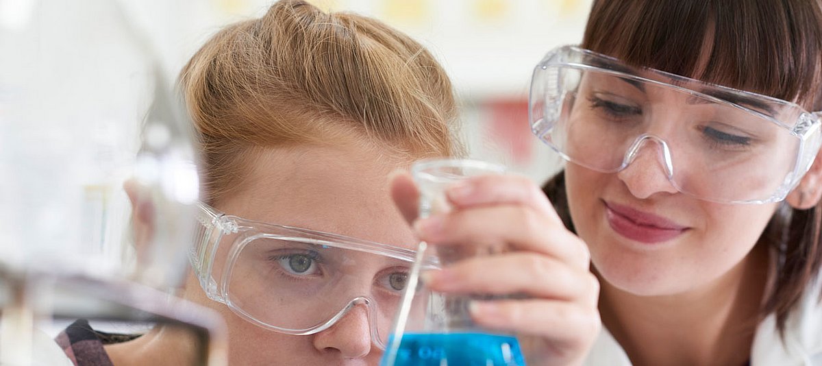 Zwei junge Frauen im Labor untersuchen eine blaue Flüssigkeit. Sie tragen Schutzbrillen.