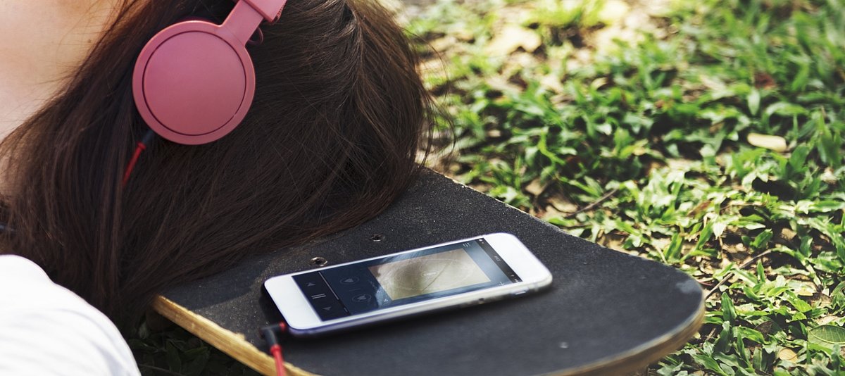 Mädchen liegt mit ihrem Kopf auf einem Skateboard und hört über Kopfhörer Musik mit ihrem Smartphone