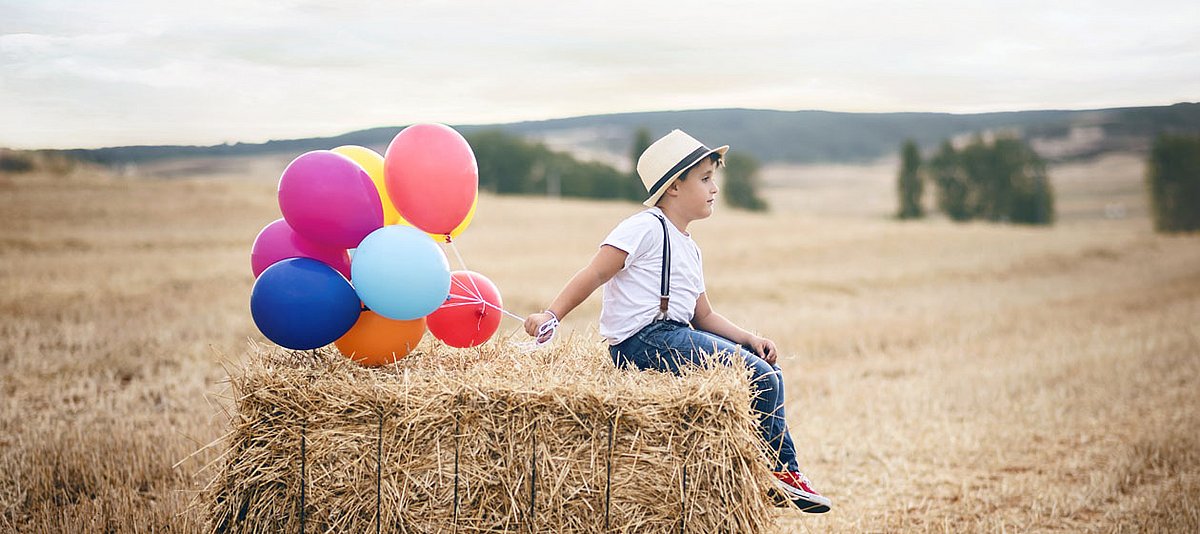 Ein Junge sitz mit einem Strohhut auf einem Strohballen und hält neun gasgefüllte Luftballons in seiner Hand.