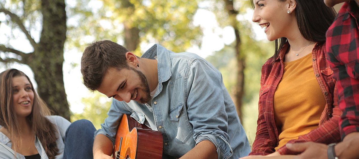 Ein junger Mann spielt Gitarre, weitere junge Menschen sitzen bei ihm und lachen