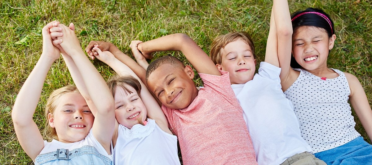 Fünf Kinder liegen mit dem Rücken auf einer Wiese, lächeln und halten sich an den Händen