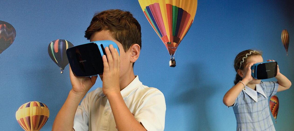 Auf dem Foto sind zwei Kinder mit VR-Brillen zu sehen. Im Hintergrund sind Heißluftballons abgebildet.