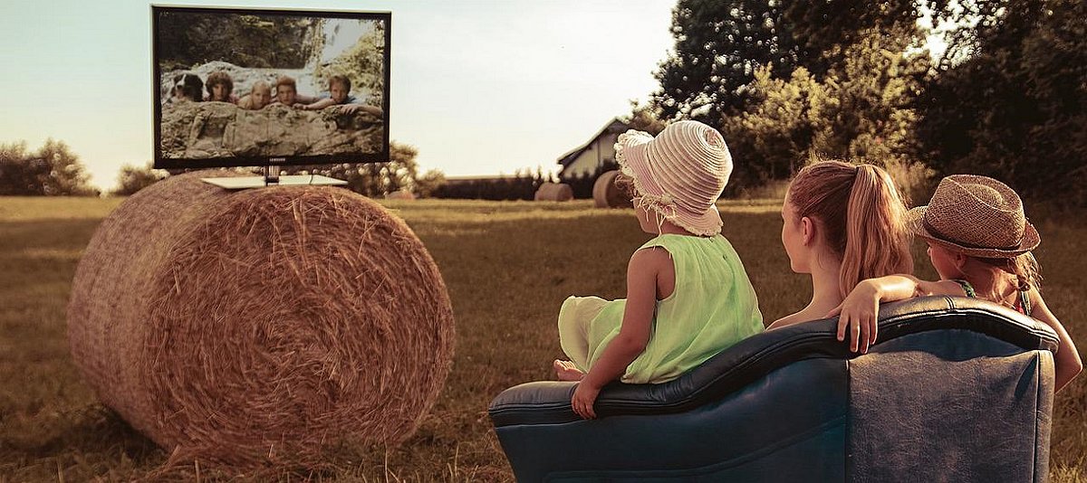 Familie schaut auf einen Fernseher und sitzt dabei auf einem Sofa, das auf einem Getreidefeld steht.