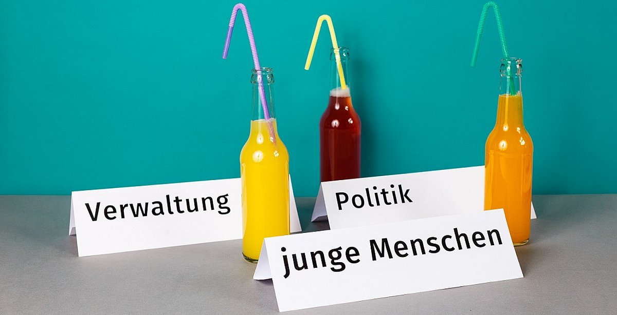 Drei Tischkarten mit den Aufschriften "Verwaltung", "Politik" und "junge Menschen" neben drei bunten Limo-Flaschen