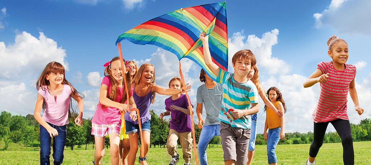 Kinder rennen über eine Wiese und lassen einen regenbogenfarbenden Drachen steigen.