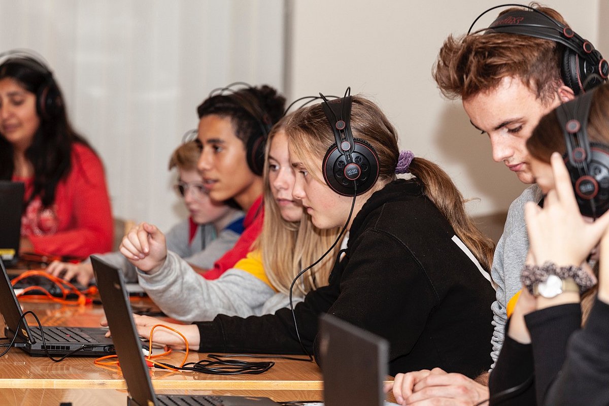 Mehrere junge Menschen sitzen mit großen Kopfhörern vor Computern
