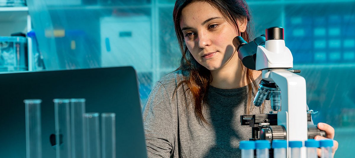 Eine junge Frau sitzt im Labor vor einem Mikroskop und schaut auf den Bildschirm eines Laptops.