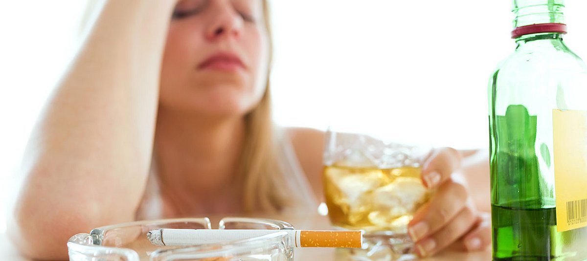 Eine Frau trinkt hochprozentigen Alkohol und sitzt sehr traurig vor der Falsche und einer brennenden Zigarette im Aschenbecher.