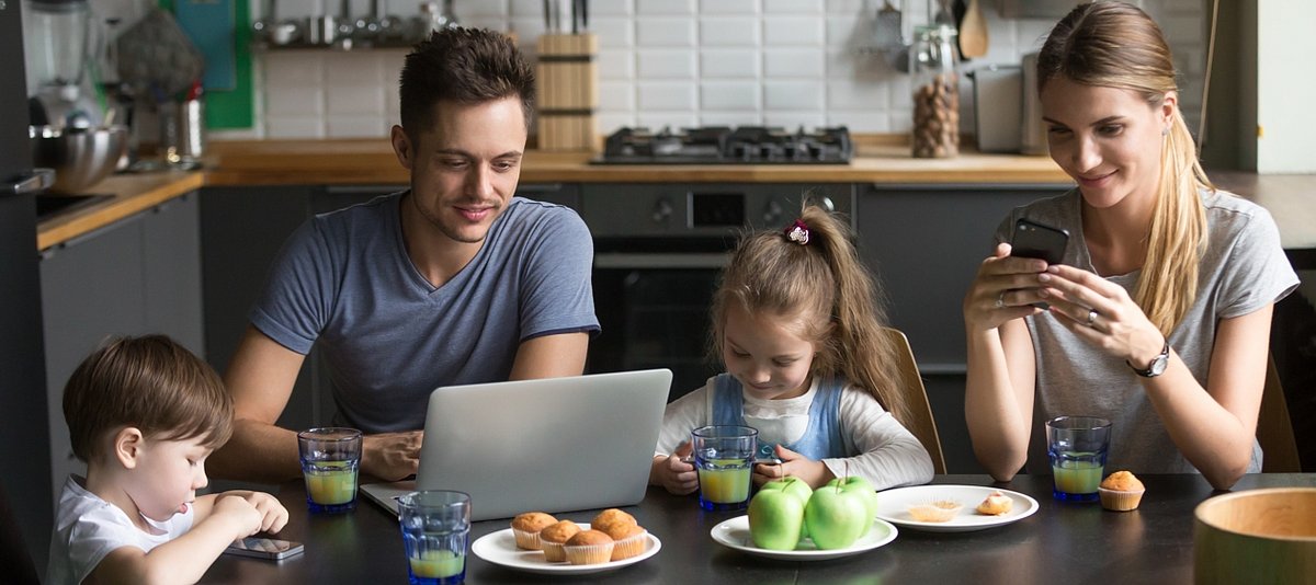Vater und Mutter sitzen mit ihren beiden Kindern am Küchentisch und alle Familienmitglieder nutzen digitale Geräte
