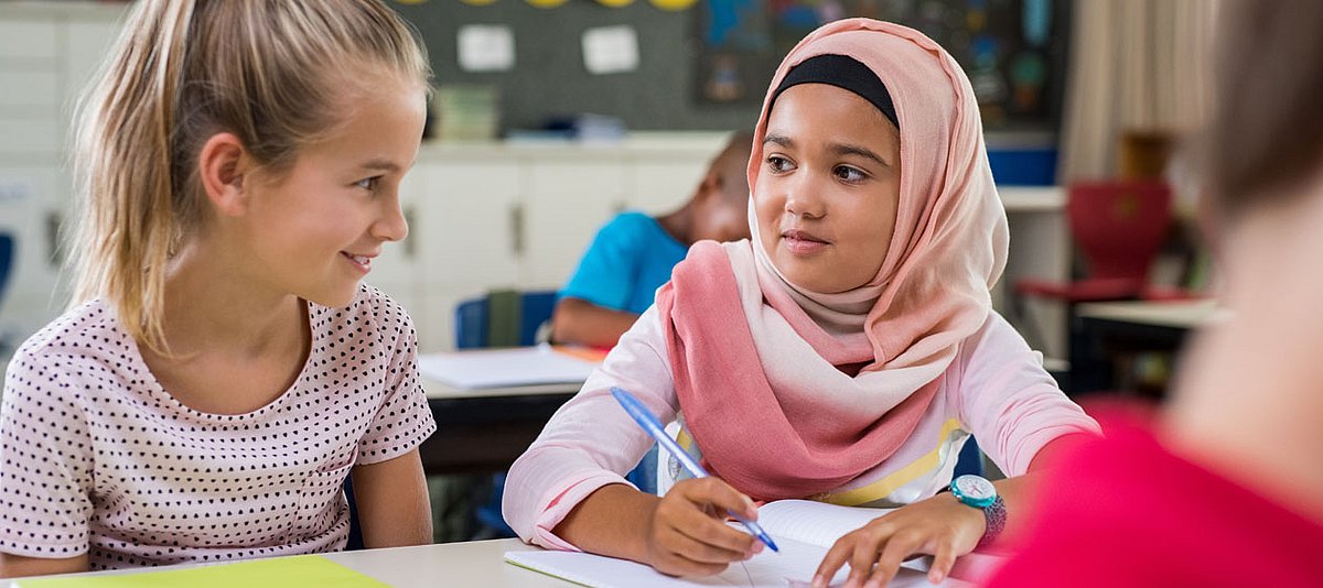 Zwei Mädchen unterschiedlicher Herkunft sitzen im Klassenzimmer nebeneinander und lächeln sich an