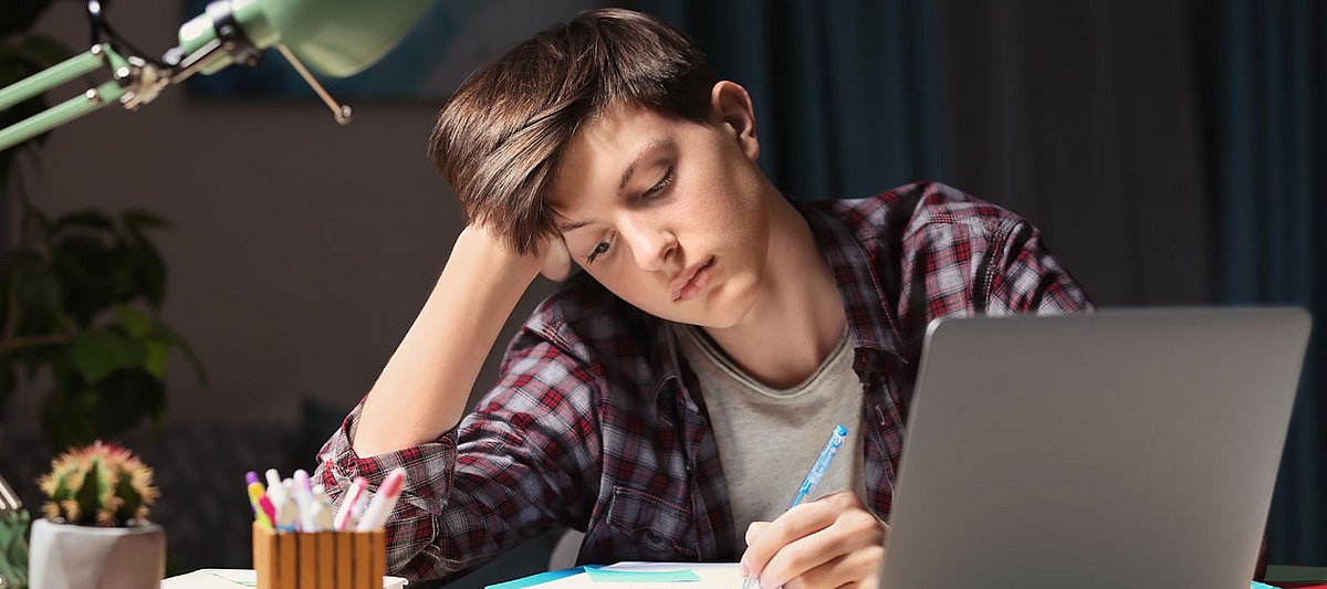 Ein Schüler sitzt unglücklich an einem Schreibtisch vor einem Laptop.