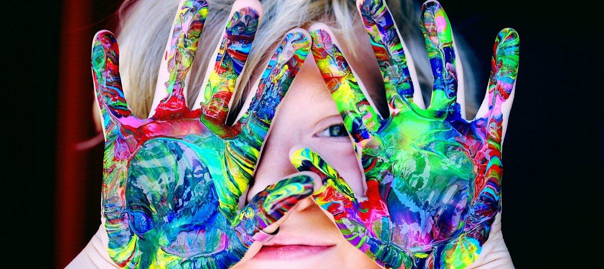 Ein Kind hält sein mit bunten Farben bedeckten Hände vors Gesicht