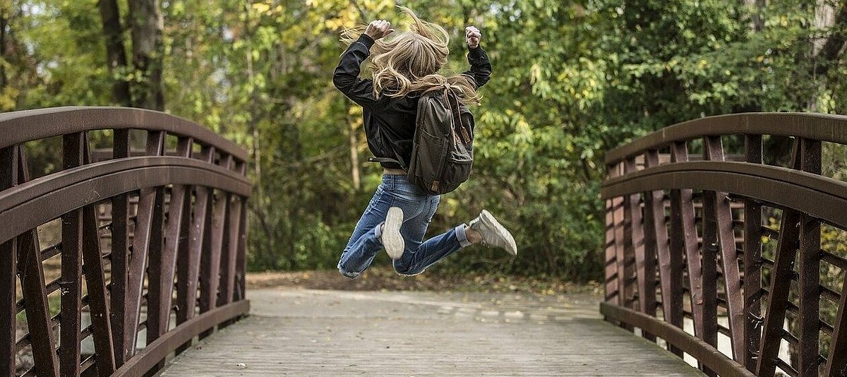 Ein blondes Mädchen hat einen Rücksack auf und macht einen Luftsprung auf einer Holzbrücke.