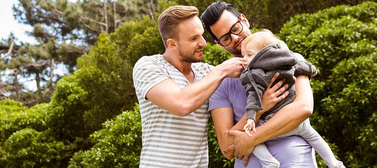 Zwei homosexuelle Väter sind im Park und einer hat ein Baby auf dem Arm, das der andere streichelt.