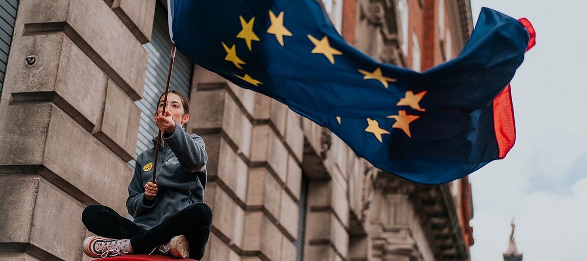 Eine junge Frau schwenkt eine große Europaflagge, im Hintergrund ist ein Gebäude zu sehen