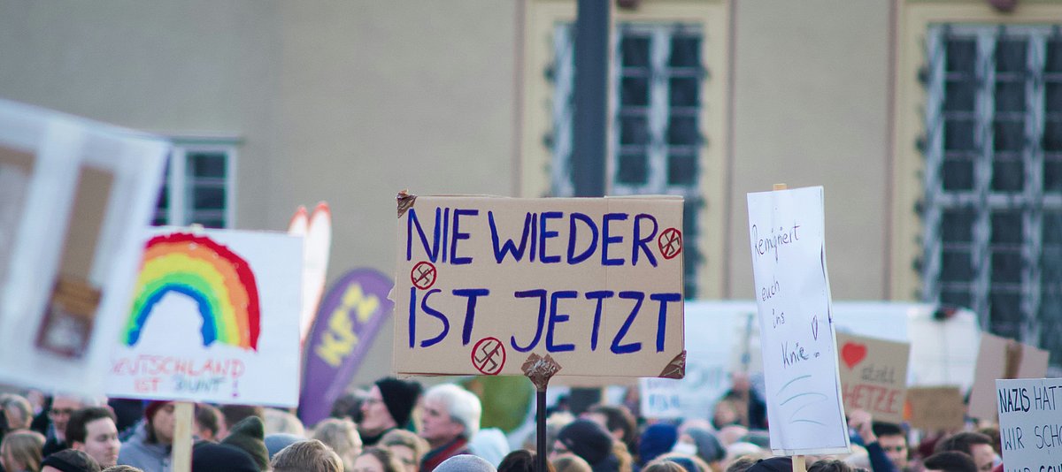 Schilder auf einer Demonstration. Im Mittelpunkt ein Schild mit der Aufschrift „Nie wieder ist jetzt“ umgeben von durchgestrichenen Hakenkreuzen. Im Hintergrund unscharf ein Schild mit einem Regenbogen und der Aufschrift „Deutschland ist bunt!“