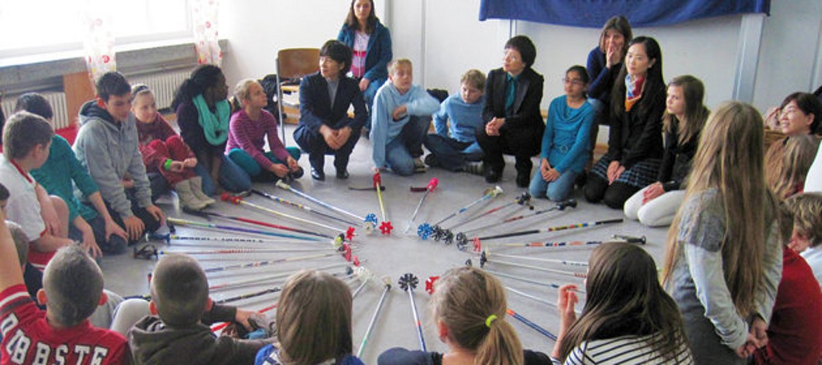 Chinesische und deutsche Kinder sitzen zusammen mit Fachkräften im Kreis auf dem Boden.