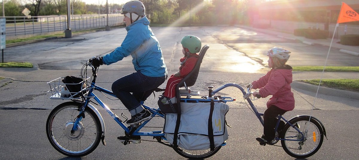 Eine erwachsene Person und zwei Kinder fahren mit Fahrradhelmen auf einem Tridem