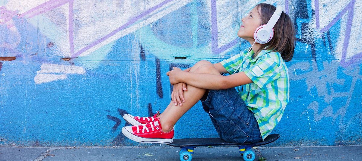 Ein Junge sitzt auf einem Skateboard vor einer Wand mit Graffitti und hört etwas mit Kopfhörern