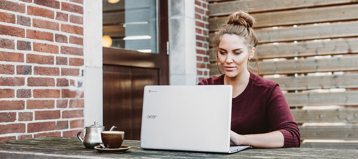 Eine junge Frau sitzt draußen und arbeitet an ihrem Laptop, auf dem Tisch steht eine Kanne und eine Tasse