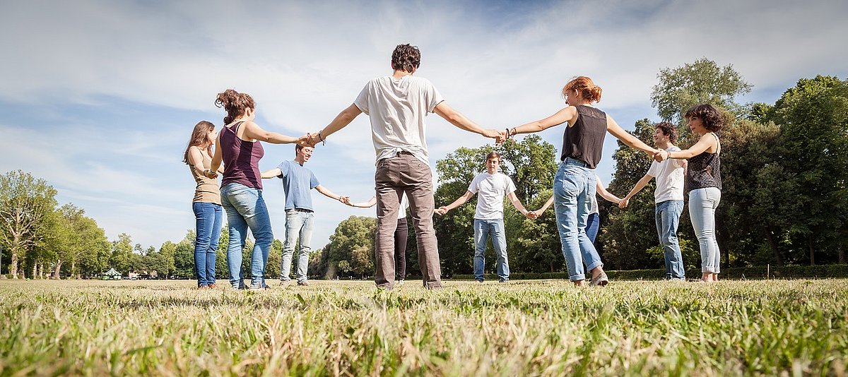 Zehn junge Erwachsene stehen im Kreis auf einer Wiese und halten sich die Hände.