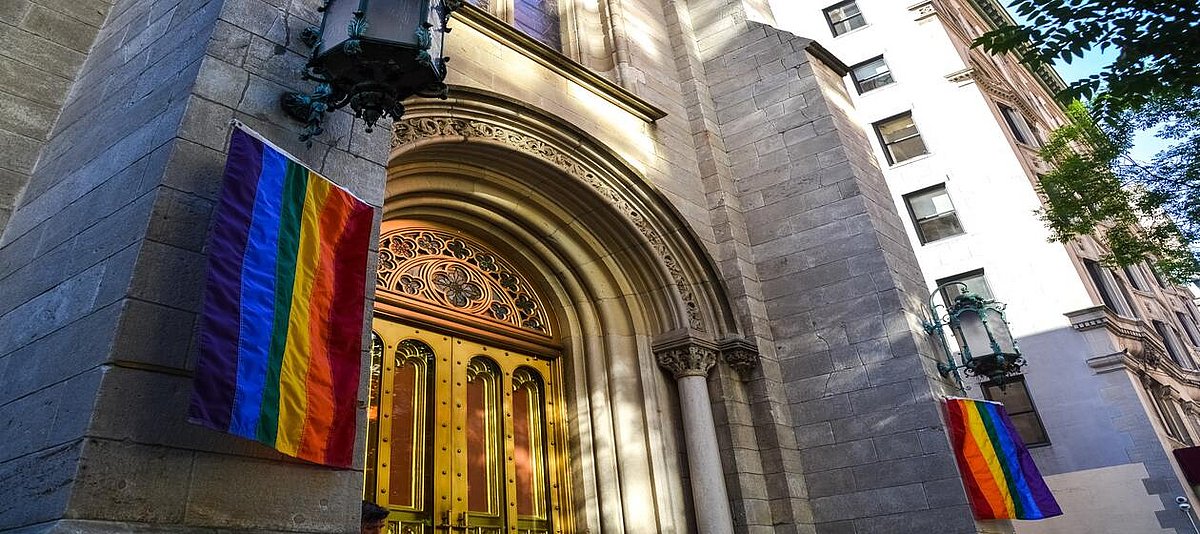 Auf dem Bild ist eine Kirchentür zu sehen, an deren Seiten zwei Regenbogenflaggen gehisst sind.