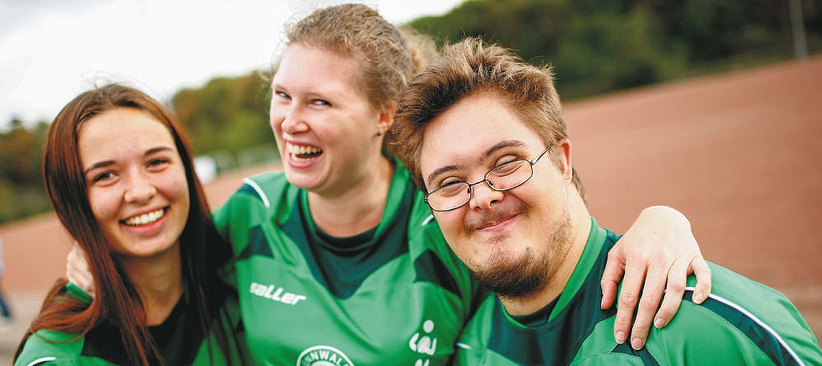 Zwei junge Frauen und ein junger Mann mit Down-Syndrom umarmen sich auf einem Sportplatz.