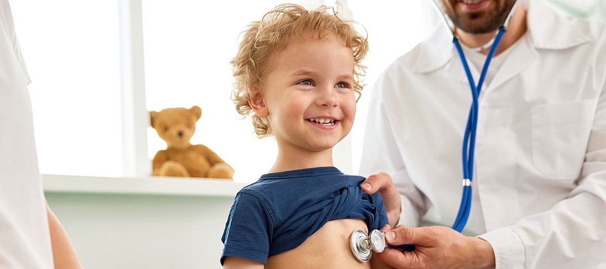 Ein Junge lächelt beim Arztbesuch, während er mit einem Stethoskop abgehört wird.