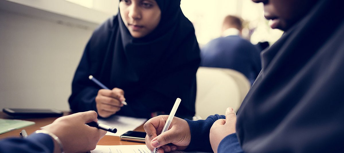 Eine Muslimin sitzt mit Klassenkameradinnen an einem Tisch im Klassenzimmer und notieren sich etwas in ihren Unterlagen.  