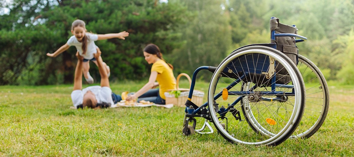 Auf einer Wieser spielen Eltern mit ihrer Tochter, im Vordergrund ist ein Rollstuhl zu sehen