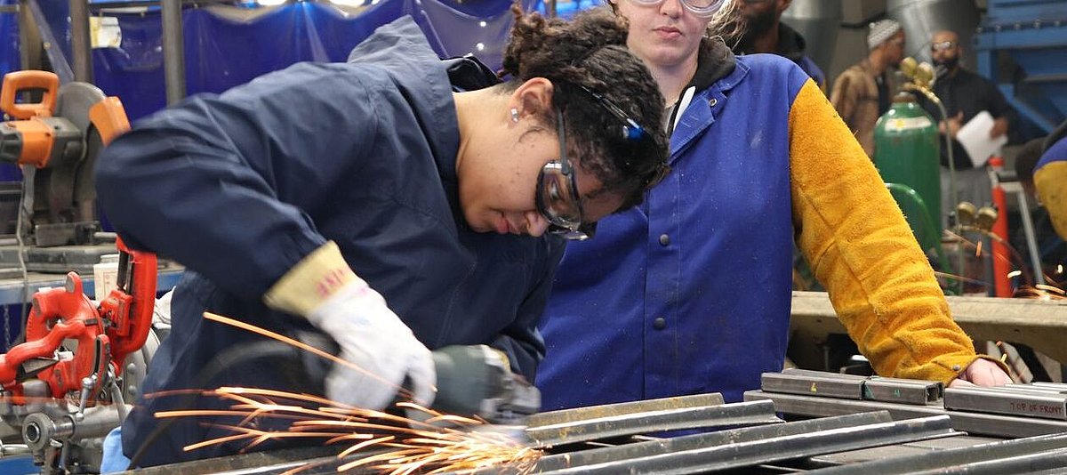 Zwei Handwerkerin im Overall schleifen in einer großen Werkstatt Metall mit einem Winkelschleifer.